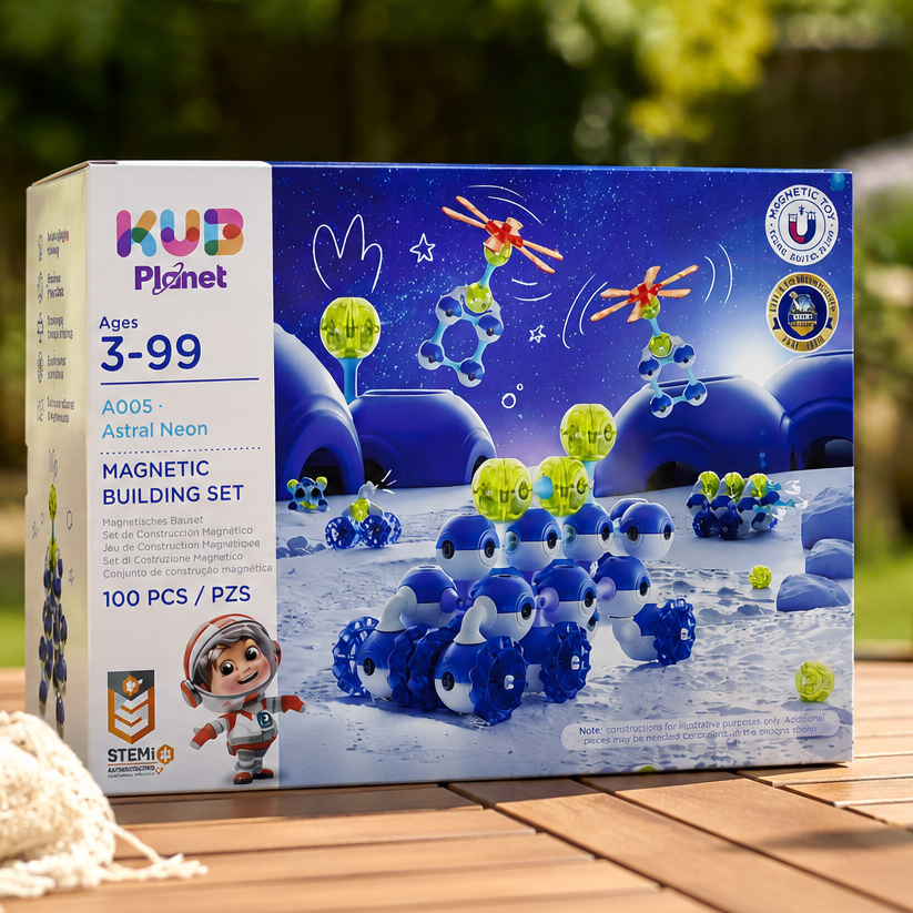A boxed toy set labeled HUB Planet Magnetic Building Set sits on a wooden outdoor table with a blurred garden background. The box is blue with space-themed graphics and product details, while the scene includes plants, a chair, and a glass on the table.